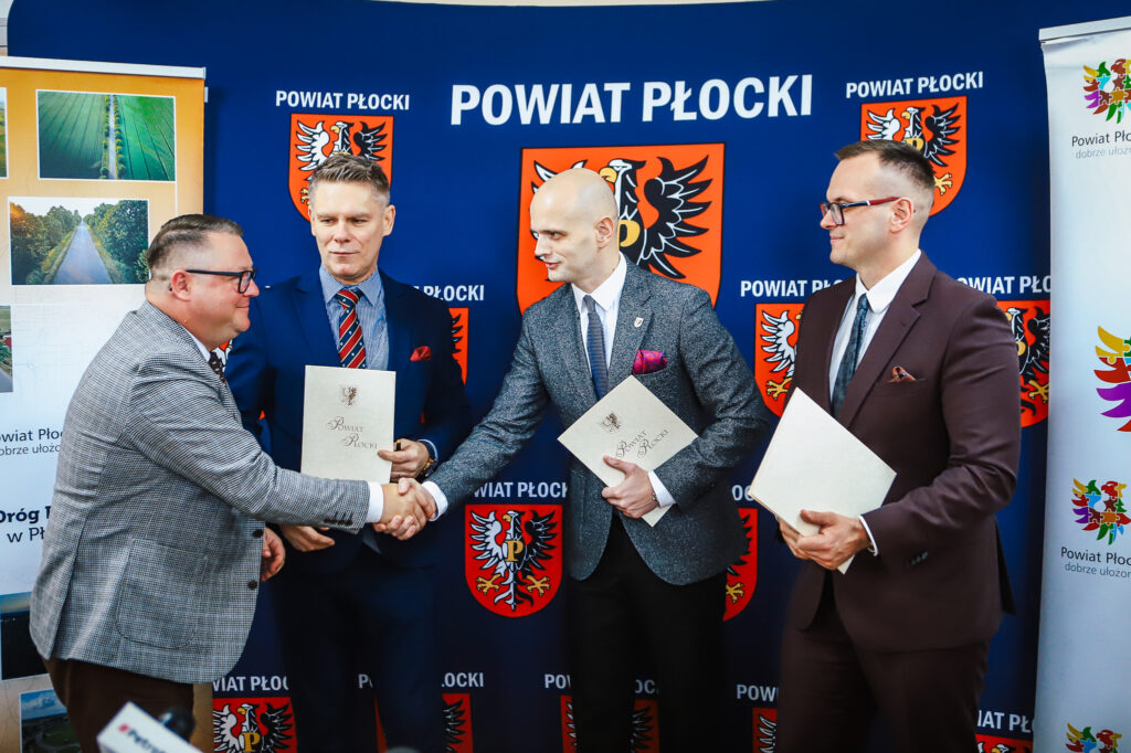 Na zdjęciu przedstawiono czterech mężczyzn w garniturach, którzy uściskiem dłoni celebrują podpisanie umowy dotyczącej budowy nowej ścieżki rowerowej w Powiecie Płockim, na tle logo Powiatu Płockiego.