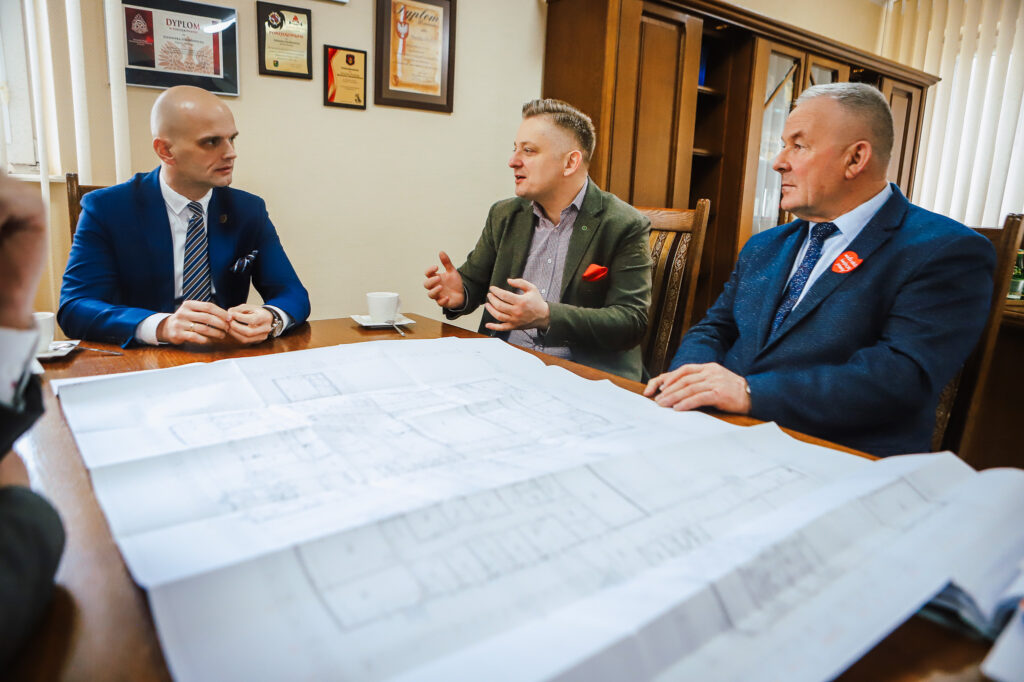 Wiceminister w Powiecie Płockim prowadzi dyskusję o bezpieczeństwie i współpracy rządu z samorządem przy stole, na którym leży plan budynku.