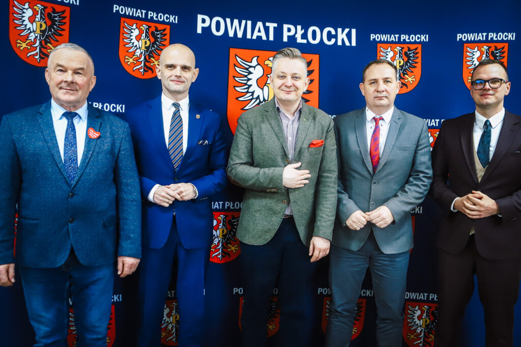 Na zdjęciu widać grupę pięciu mężczyzn stojących obok siebie przed tłem z herbem Powiatu Płockiego, podczas wizyty Wiceministra w Powiecie Płockim, podkreślając współpracę rządu z samorządem.