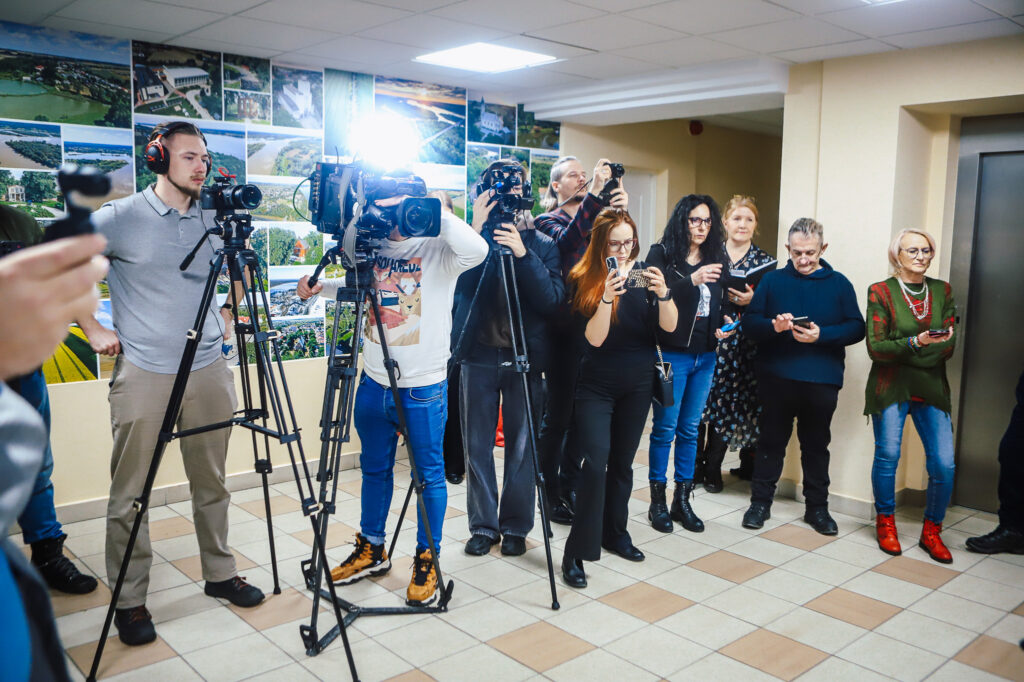 Grupa dziennikarzy i fotografów z aparatami oraz smartfonami uczestniczy w wydarzeniu związanym z wizytą Wiceministra w Powiecie Płockim, promującym bezpieczeństwo i współpracę rządu z samorządem.