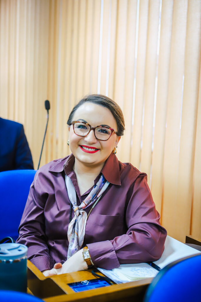 Na zdjęciu widoczna jest uśmiechnięta kobieta w fioletowej koszuli oraz z apaszką, siedząca w sali obrad podczas XVIII Sesji Rady Powiatu w Płocku.