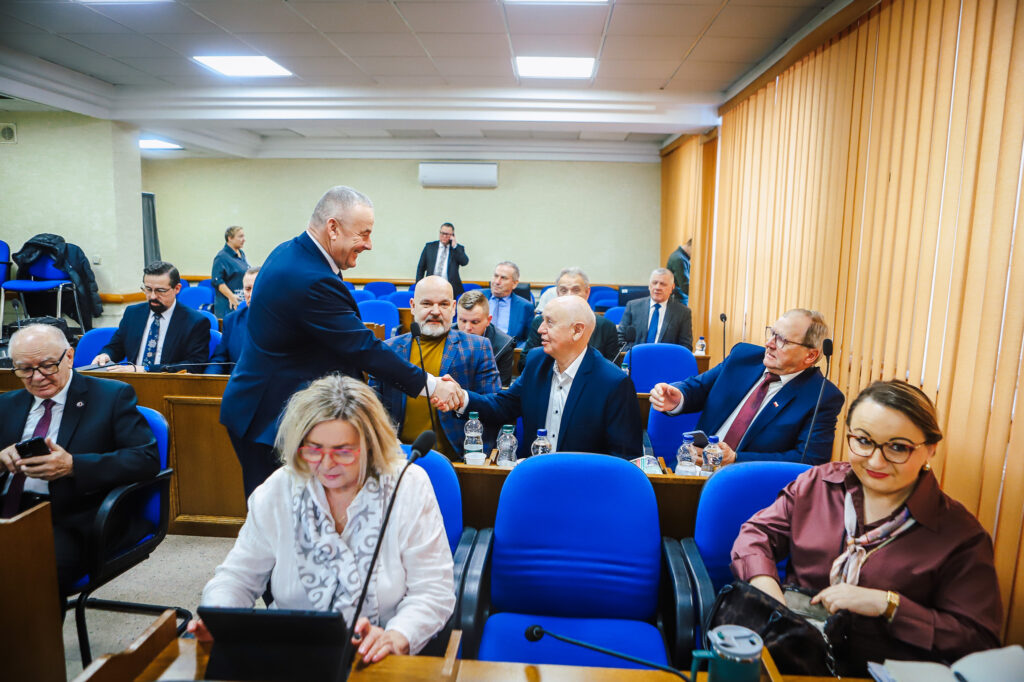 Na zdjęciu przedstawiono uczestników XVIII Sesji Rady Powiatu w Płocku, którzy w atmosferze współpracy wymieniają uściski dłoni i dyskutują w sali obrad.