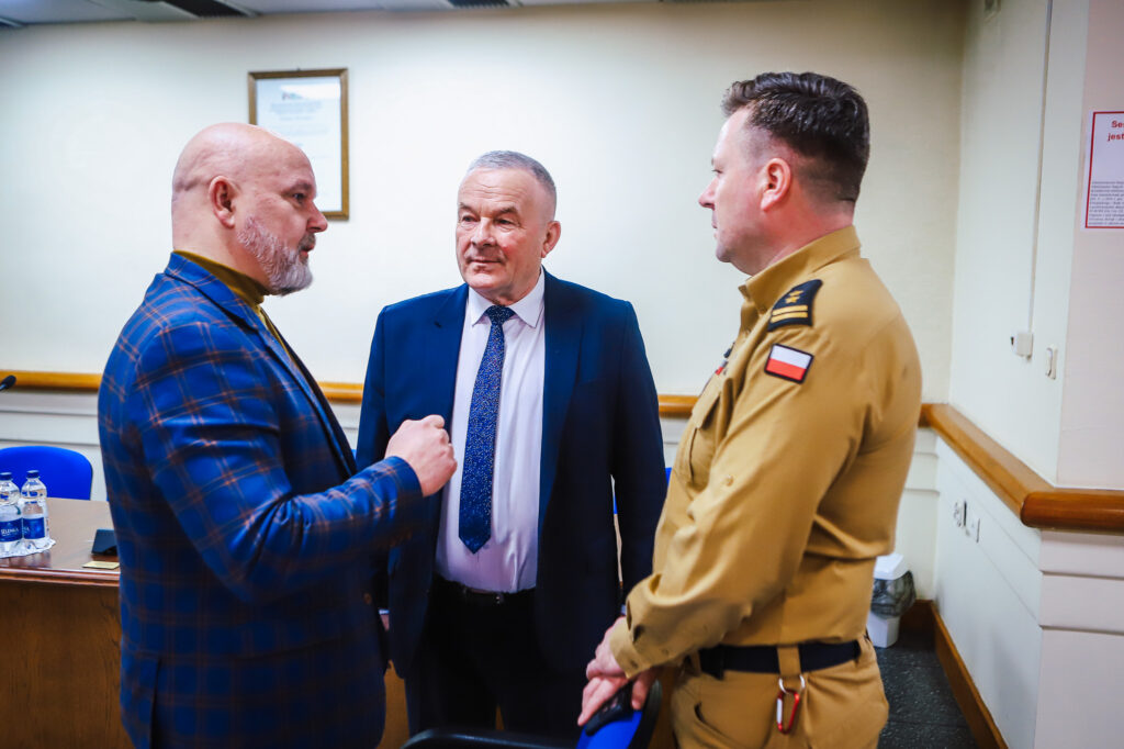 Na zdjęciu widać trzech mężczyzn rozmawiających w sali obrad podczas XVIII Sesji Rady Powiatu w Płocku, podkreślających znaczenie jednomyślnych decyzji oraz wsparcia społecznego.