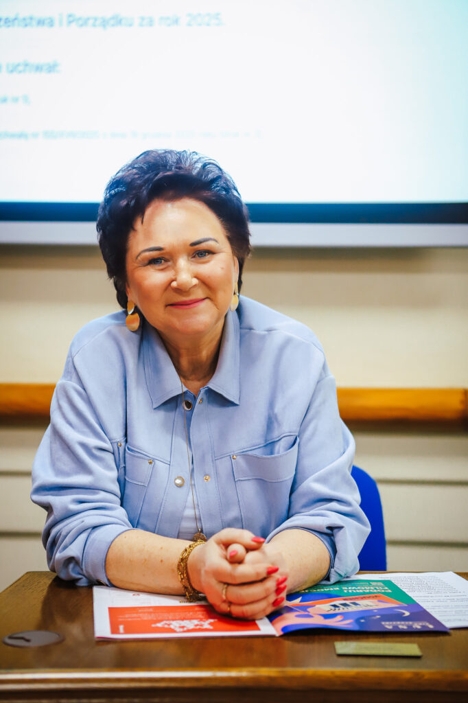 Uśmiechająca się kobieta w niebieskiej bluzie siedzi przy stole, podczas XVIII Sesji Rady Powiatu w Płocku.