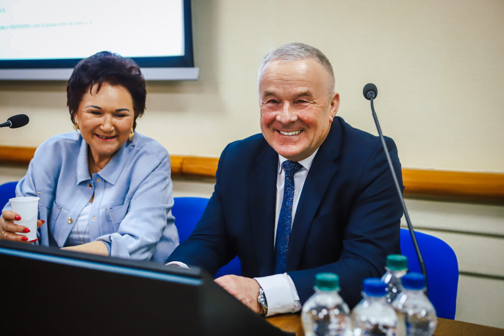 Na zdjęciu widać uśmiechniętą parę ludzi podczas XVIII Sesji Rady Powiatu w Płocku, którzy uczestniczą w dyskusji dotyczącej jednomyślnych decyzji i wsparcia społecznego.