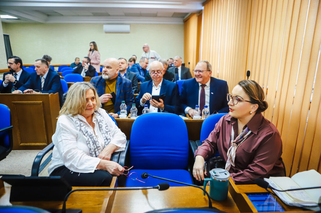 Na zdjęciu widać uczestników XVIII Sesji Rady Powiatu w Płocku, którzy dyskutują między sobą w sali obrad z niebieskimi krzesłami i drewnianymi elementami wnętrza.