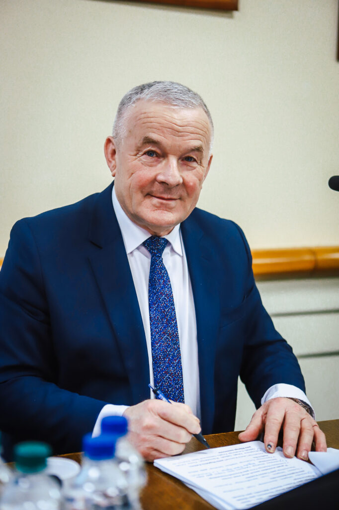 Na zdjęciu widać uśmiechniętego mężczyznę w garniturze, który pisze na spotkaniu Rady Powiatu w Płocku.