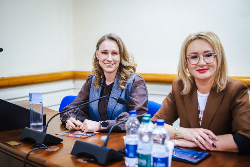 Na zdjęciu widać dwie uśmiechnięte kobiety siedzące przy stole w trakcie XVIII Sesji Rady Powiatu w Płocku.