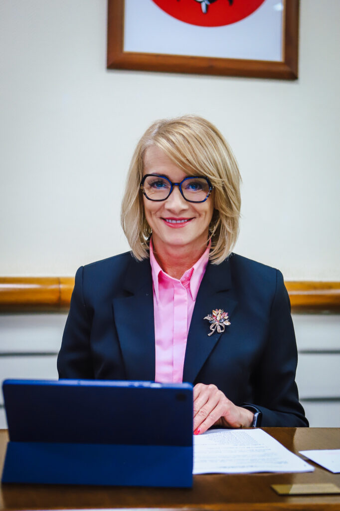 Na zdjęciu przedstawiona jest kobieta w eleganckim stroju, z uśmiechem, siedząca przy stole z tabletem i dokumentami, podczas XVIII Sesji Rady Powiatu w Płocku.