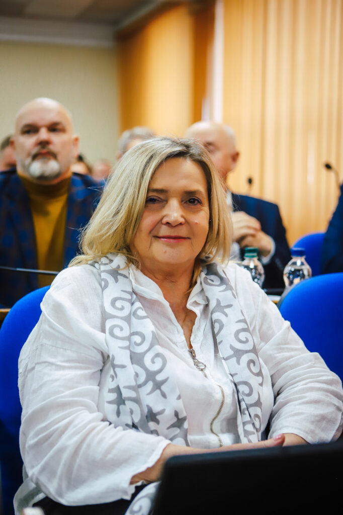 Kobieta w białej bluzce z szalem siedzi w sali obrad, uśmiechając się i uczestnicząc w XVIII Sesji Rady Powiatu w Płocku.