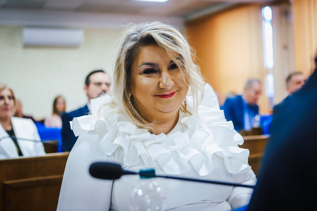 Na zdjęciu widać uśmiechniętą kobietę w białej bluzce z falbanami, uczestniczącą w XVIII Sesji Rady Powiatu w Płocku.