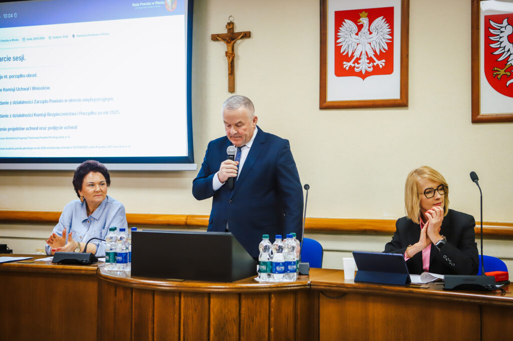 Na zdjęciu przedstawiono przebieg XVIII Sesji Rady Powiatu w Płocku, podczas której omawiane są istotne tematy dotyczące wsparcia społecznego, a w centrum uwagi znajduje się mężczyzna mówiący do zgromadzonych członków rady.