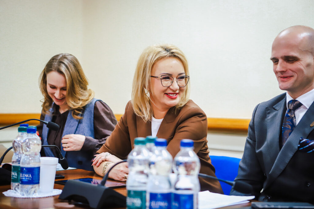 Na zdjęciu widać uśmiechniętych uczestników XVIII Sesji Rady Powiatu w Płocku, siedzących przy stole w formalnej sali obrad.