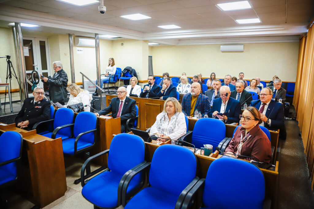 Zdjęcie przedstawia uczestników XVIII Sesji Rady Powiatu w Płocku, którzy z zaangażowaniem dyskutują w nowoczesnej sali obrad.