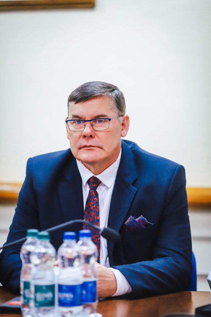 Na zdjęciu widać mężczyznę w garniturze, siedzącego przy stole z butelkami wody, podczas XVIII Sesji Rady Powiatu w Płocku, gdzie omawiane są ważne tematy społeczne.