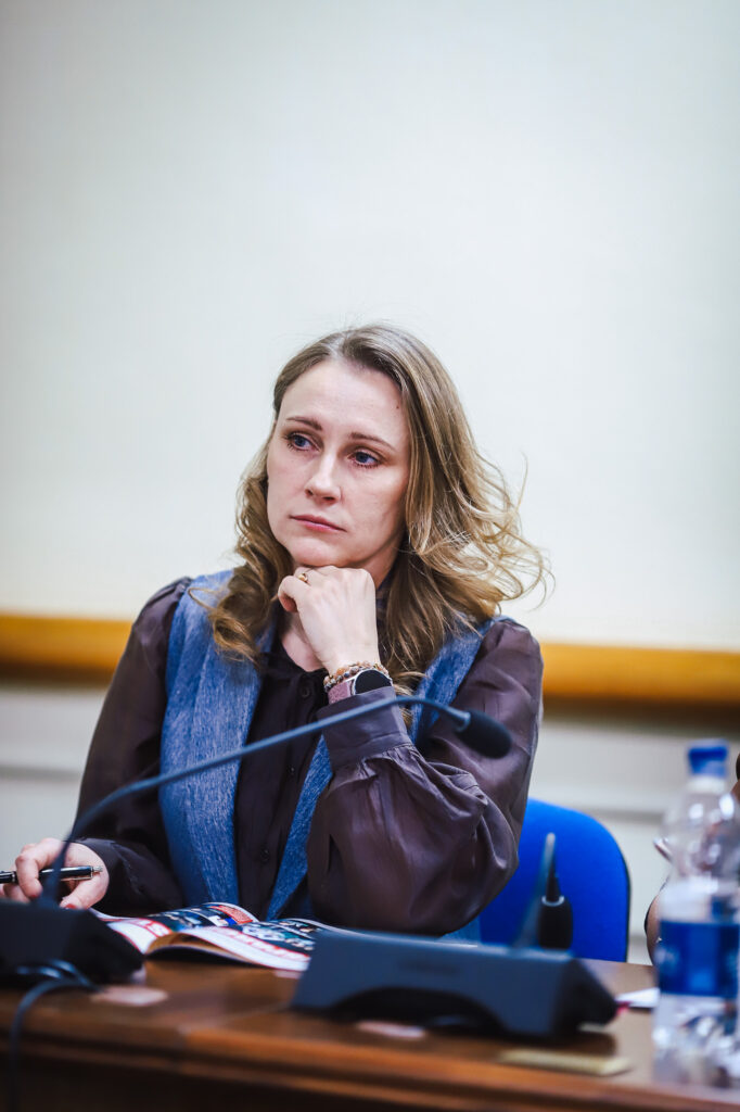 Na zdjęciu przedstawiona jest kobietę w trakcie XVIII Sesji Rady Powiatu w Płocku, która z zaangażowaniem słucha dyskusji, trzymając długopis i przyglądając się papierom na stole.