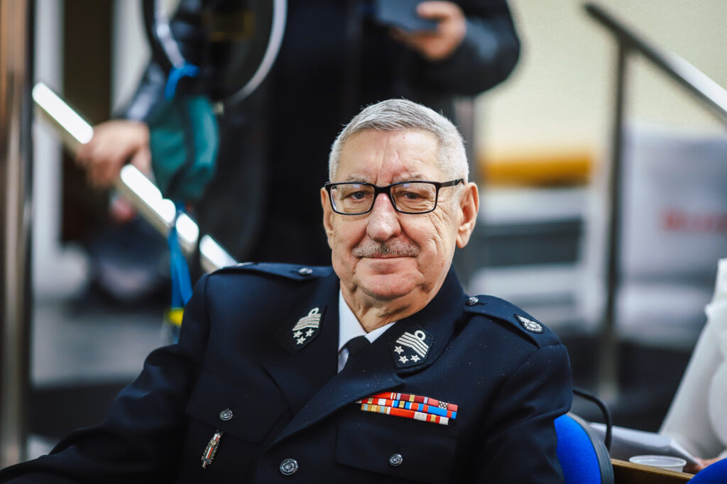 Na zdjęciu widać uśmiechniętego mężczyznę w mundurze, uczestniczącego w XVIII Sesji Rady Powiatu w Płocku.