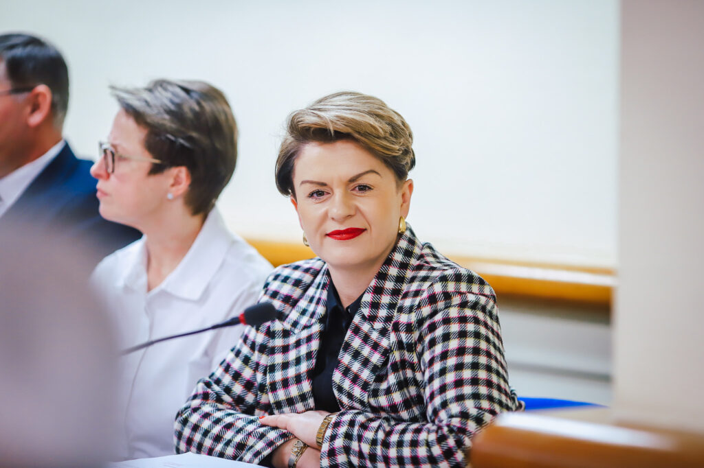 Uczestniczka XVIII Sesji Rady Powiatu w Płocku uśmiecha się znad stołu, z mikrofonem w tle, reprezentując ważne decyzje i wsparcie społeczne.