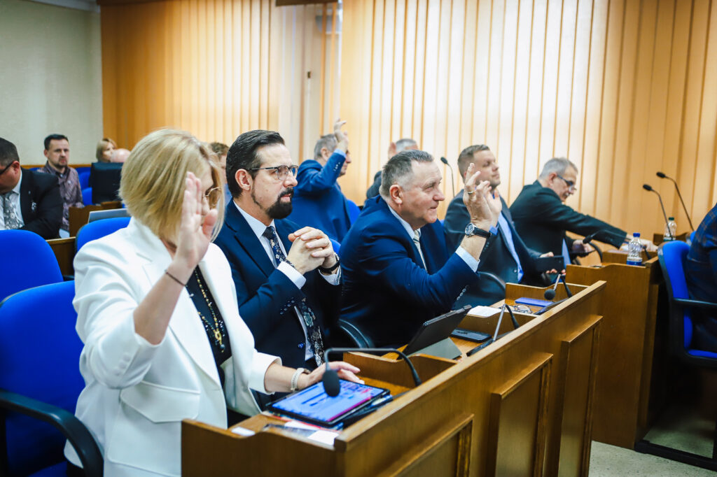 Na zdjęciu widoczna jest grupa osób w sali obrad, podczas XVIII Sesji Rady Powiatu w Płocku, gdzie członkowie podnoszą ręce w celu głosowania.