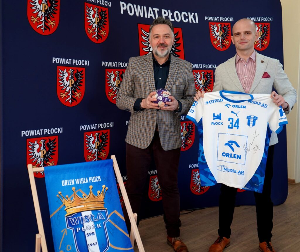Na zdjęciu dwóch mężczyzn prezentuje podpisaną koszulkę i piłkę, stojąc przed tłem z herbem Powiatu Płockiego, w kontekście wsparcia Wielkiej Orkiestry Świątecznej Pomocy.