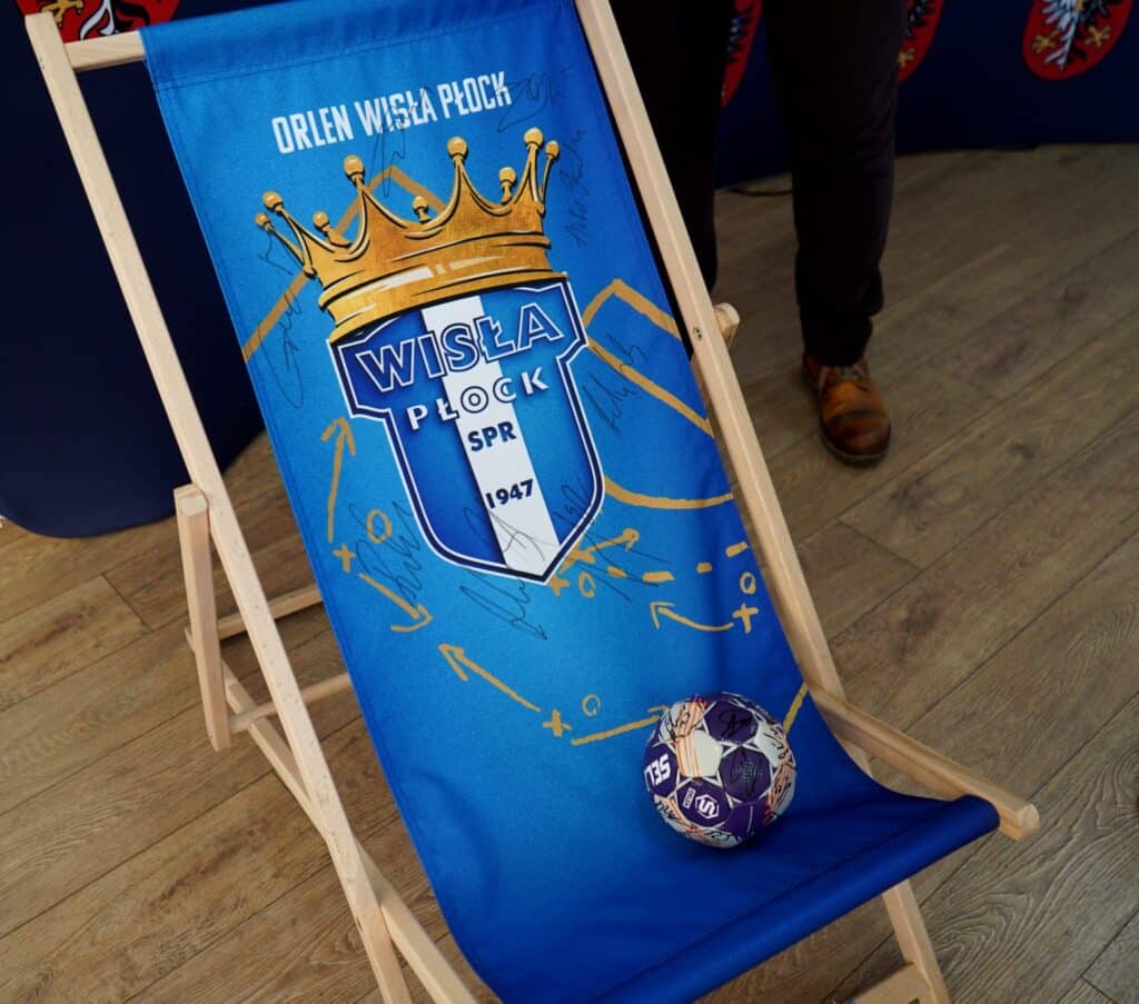 Na zdjęciu znajduje się leżak z logo klubu Orlen Wisła Płock oraz piłka ręczna ozdobiona autografami zawodników.