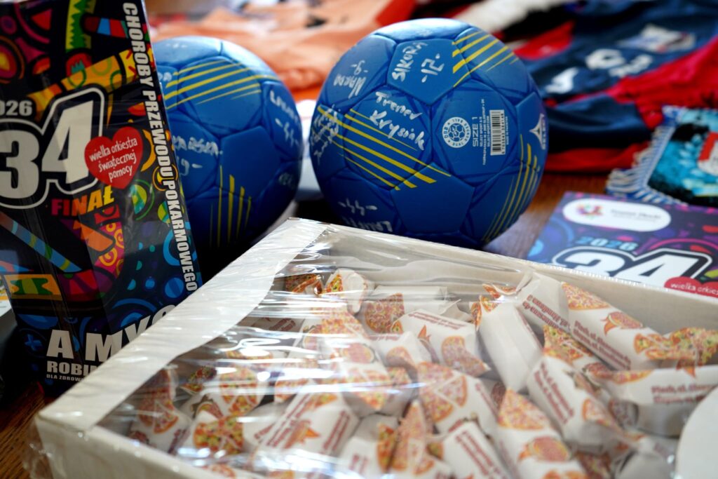 Na zdjęciu widać dwa niebieskie piłki ręczne z podpisami, pudełko cukierków oraz materiały związane z 34. finałem Wielkiej Orkiestry Świątecznej Pomocy.