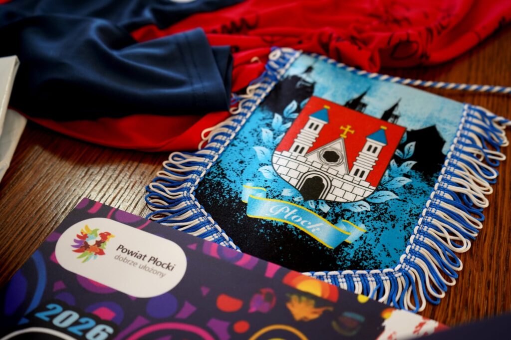 Na zdjęciu znajduje się stół z różnymi przedmiotami, wśród których wyróżnia się herb Płocka w kolorze niebieskim oraz materiały promocyjne dotyczące powiatu płockiego.