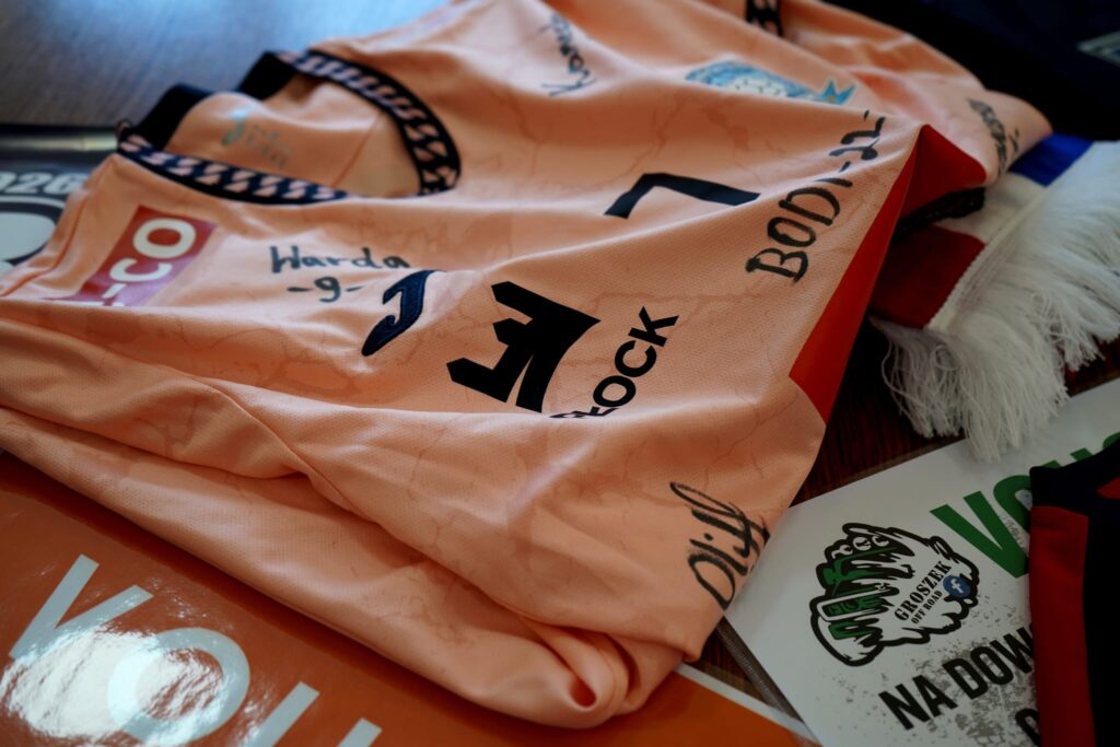 Na zdjęciu znajdują się pomarańczowe koszulki sportowe leżące na stole, obok których leżą materiały promocyjne związane ze sportem.