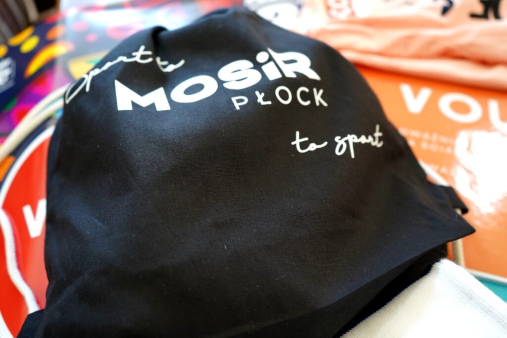 Na zdjęciu znajduje się czarna torba z napisem "MOSiR PŁOCK to sport" w otoczeniu kolorowych ubrań i logo.
