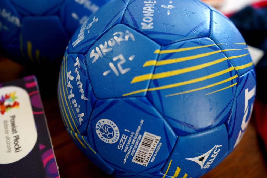 Na zdjęciu widoczna jest niebieska piłka ręczna z białymi autografami i napisami, leżąca na drewnianym stole obok kolorowej teczki.