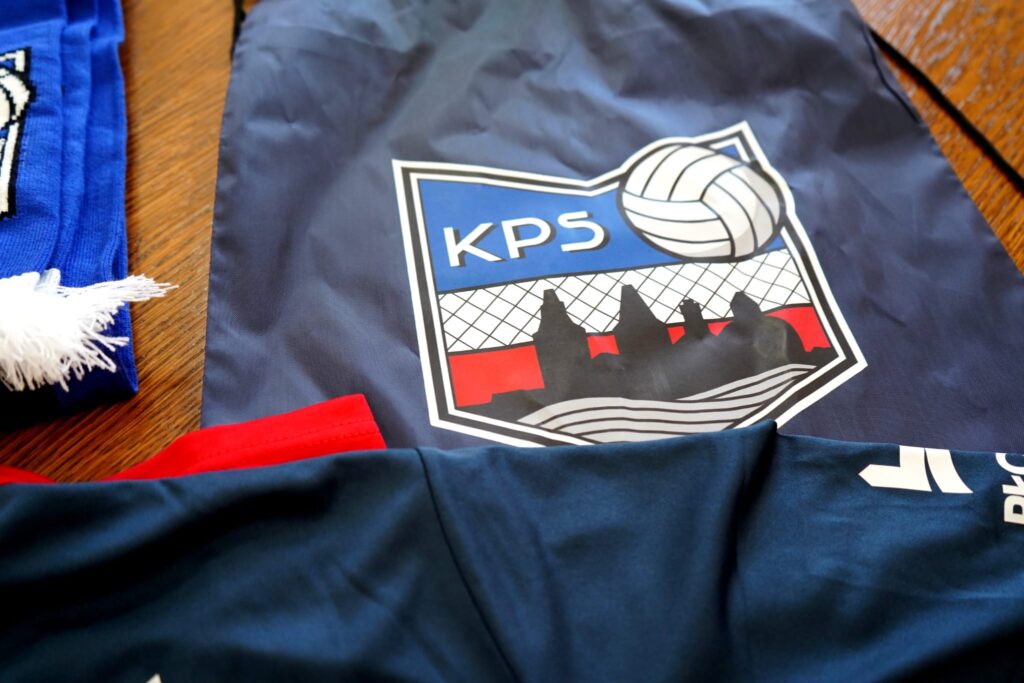 Na zdjęciu widać torbę sportową z logo klubu KPS oraz fragment kolorowego stroju piłkarskiego w odcieniach niebieskiego i czerwonego.
