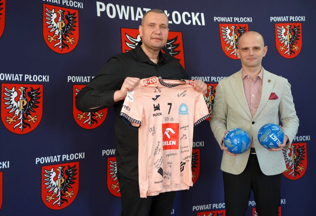 Na zdjęciu dwóch mężczyzn trzymających koszulkę podpisaną przez zawodników oraz piłki, z tłem z herbami Powiatu Płockiego, podczas akcji charytatywnej na rzecz Wielkiej Orkiestry Świątecznej Pomocy.