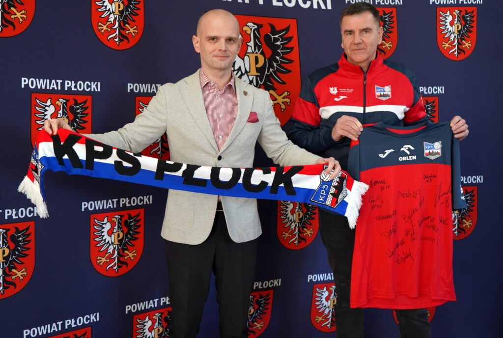 Na zdjęciu dwóch mężczyzn trzymających szalik KPS Płock oraz podkoszulek z podpisami, na tle muralu z herbem powiatu płockiego.