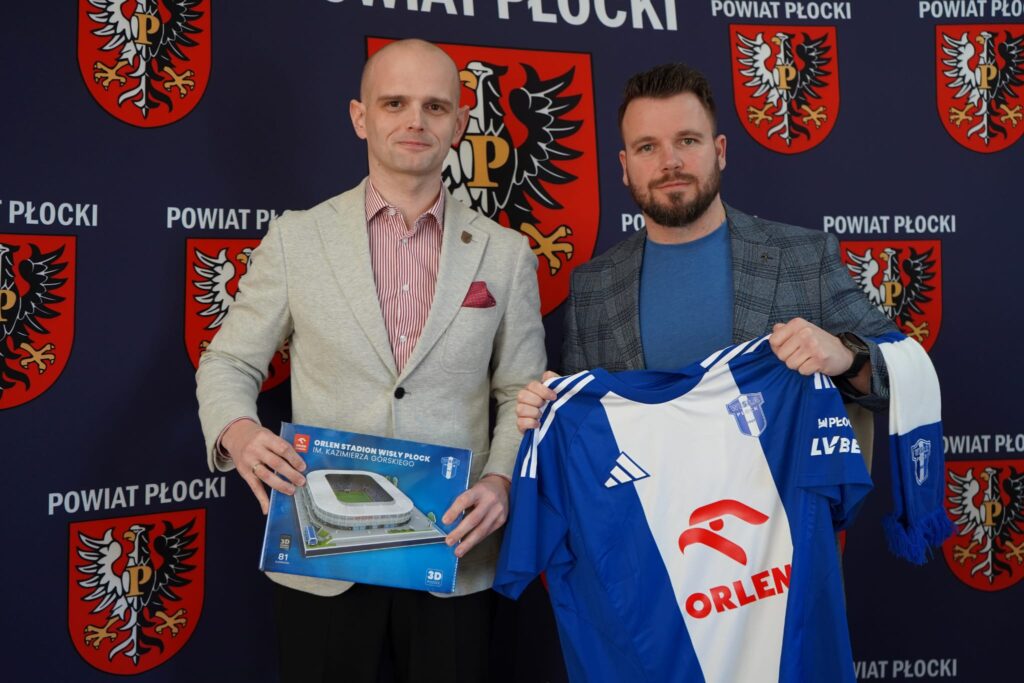 Na zdjęciu dwóch mężczyzn stoi przed tłem z herbem powiatu płockiego, trzymając makietę stadionu oraz koszulkę piłkarską, promując akcję wsparcia dla Wielkiej Orkiestry Świątecznej Pomocy.