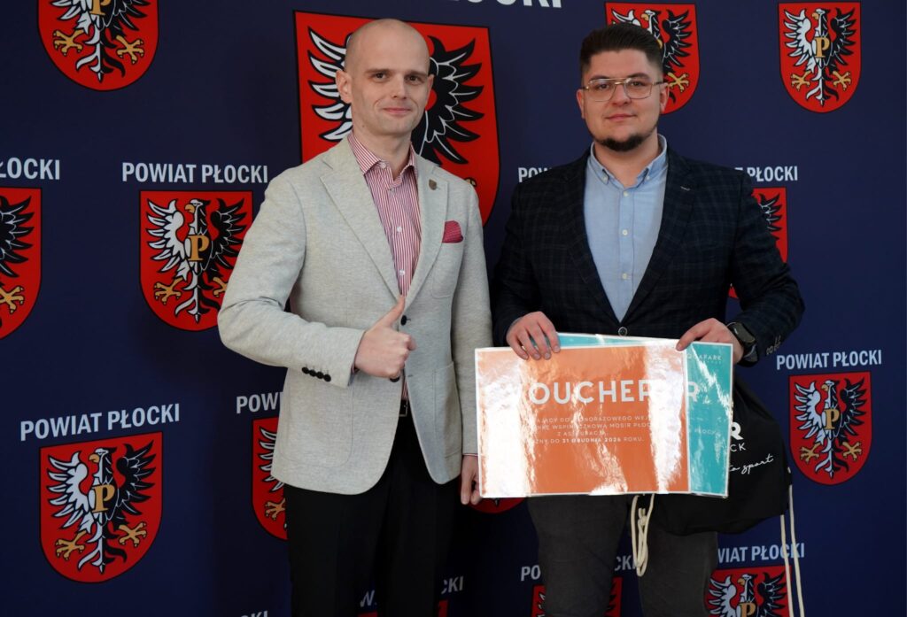 Na zdjęciu dwóch mężczyzn, jeden z nich wskazuje kciukiem w górę, trzymając voucher, w tle widoczny jest baner z herbami Powiatu Płockiego.