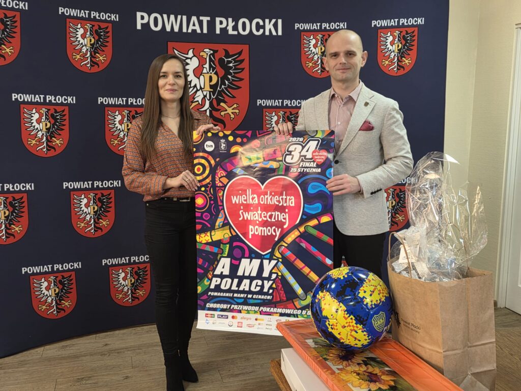 Na zdjęciu widoczni są dwie osoby trzymające plakat 34. Finału Wielkiej Orkiestry Świątecznej Pomocy, z logo powiatu płockiego w tle.
