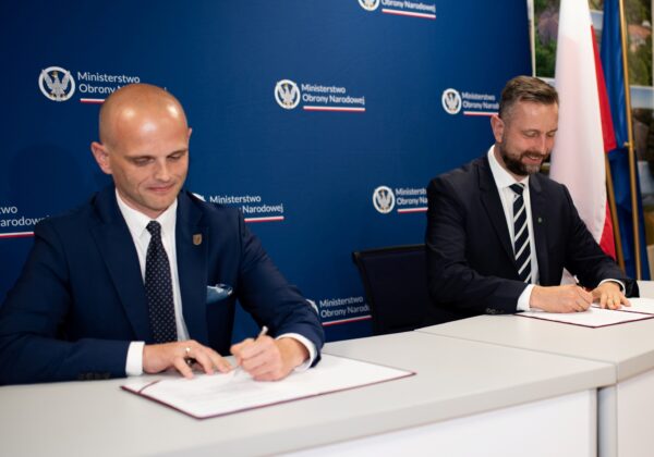 Dwóch mężczyzn w garniturach podpisuje dokumenty przy stole podczas plebiscytu „Z Tumskiego Wzgórza”, na tle niebieskiej ścianki z logo Ministerstwa Obrony Narodowej i flag.