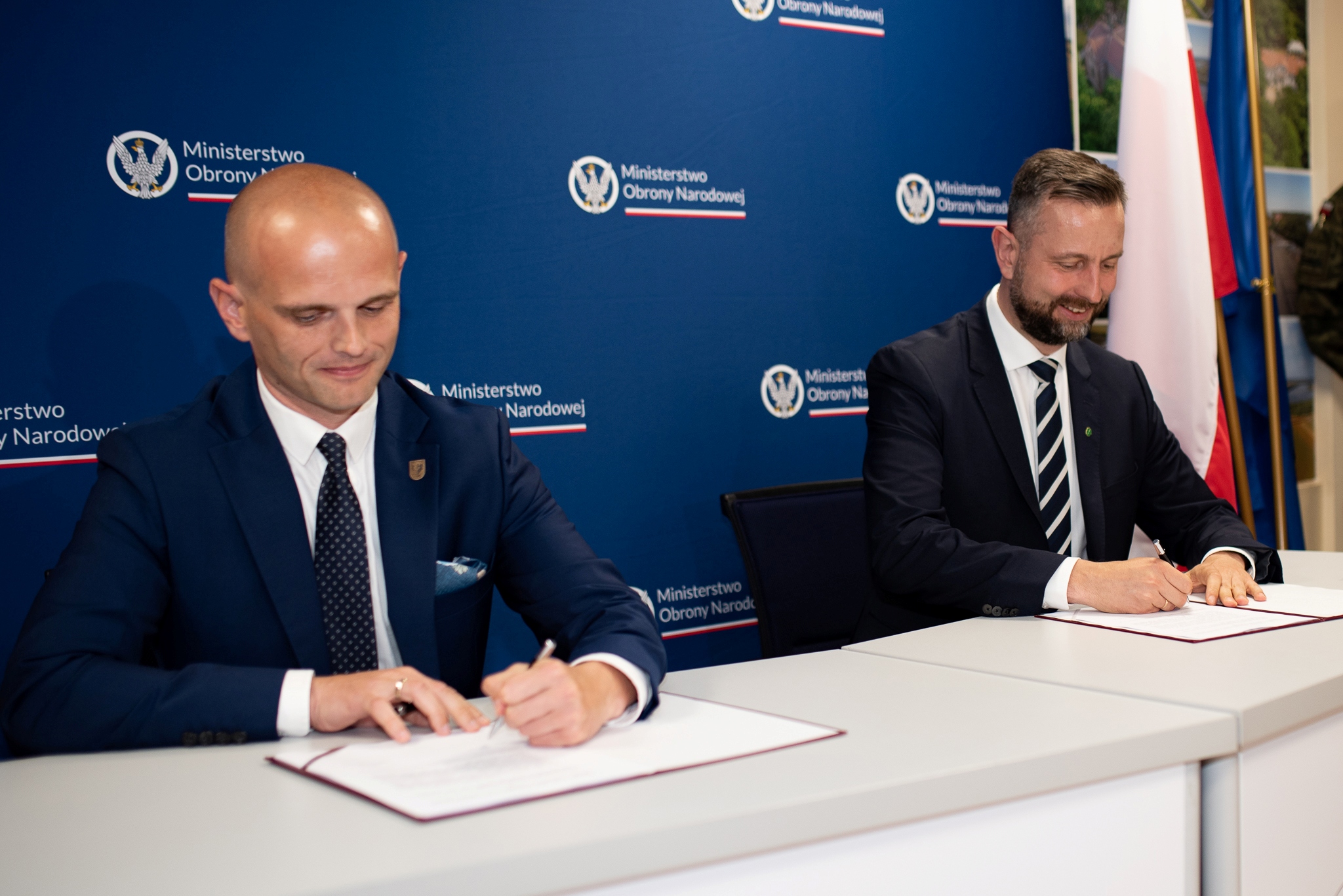 Dwóch mężczyzn w garniturach podpisuje dokumenty przy stole podczas plebiscytu „Z Tumskiego Wzgórza”, na tle niebieskiej ścianki z logo Ministerstwa Obrony Narodowej i flag.