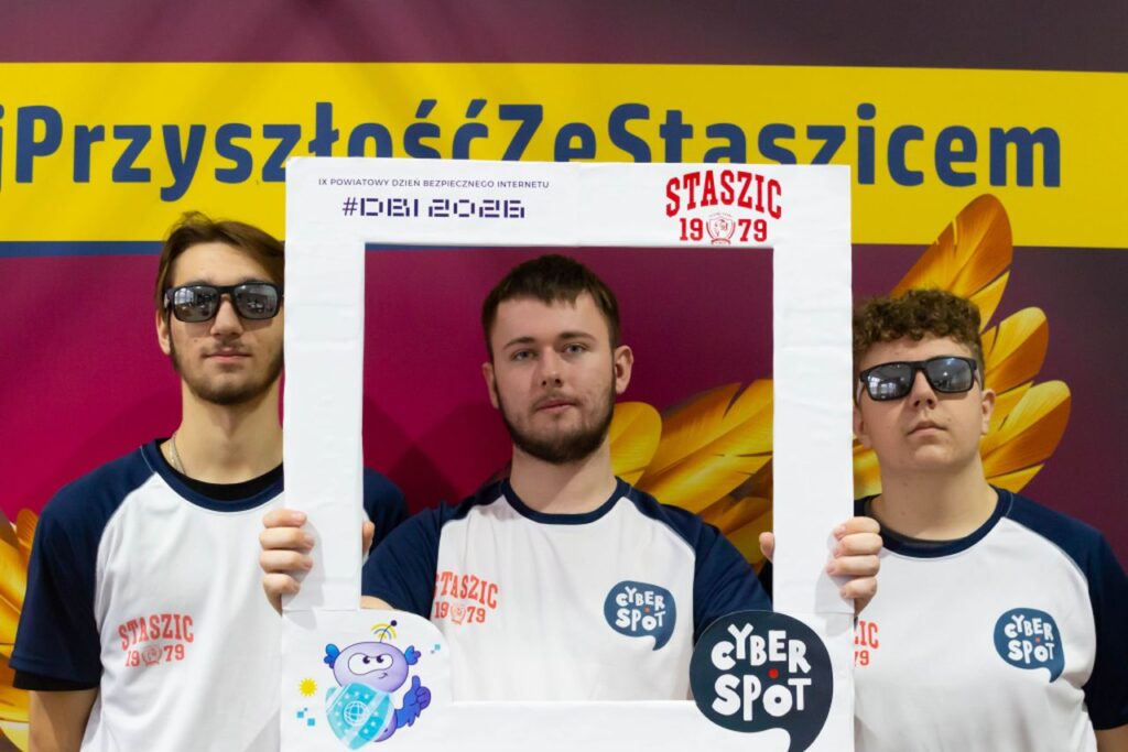 Trzech młodych mężczyzn w koszulkach z logo Cyber Spot trzyma przed sobą białą ramkę zdjęciową, stojąc na tle kolorowego baneru IX Powiatowego Dnia Bezpiecznego Internetu 2026 w Zespole Szkół im. Stanisława Staszica w Gąbinie.