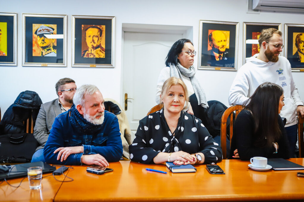 Grupa samorządowców Powiatu Płockiego i lokalnych mediów siedzi przy długim stole podczas noworocznego śniadania prasowego; na pierwszym planie kobieta w czarno-białej bluzce w groszki, a w tle widoczne ramkowe portrety na ścianie.