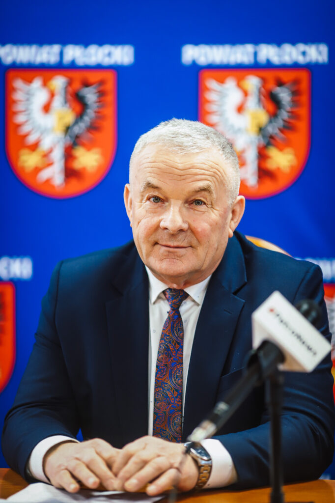 Mężczyzna w garniturze siedzi przy stole z mikrofonem podczas noworocznego śniadania prasowego Powiatu Płockiego, na niebieskim tle z herbem powiatu.