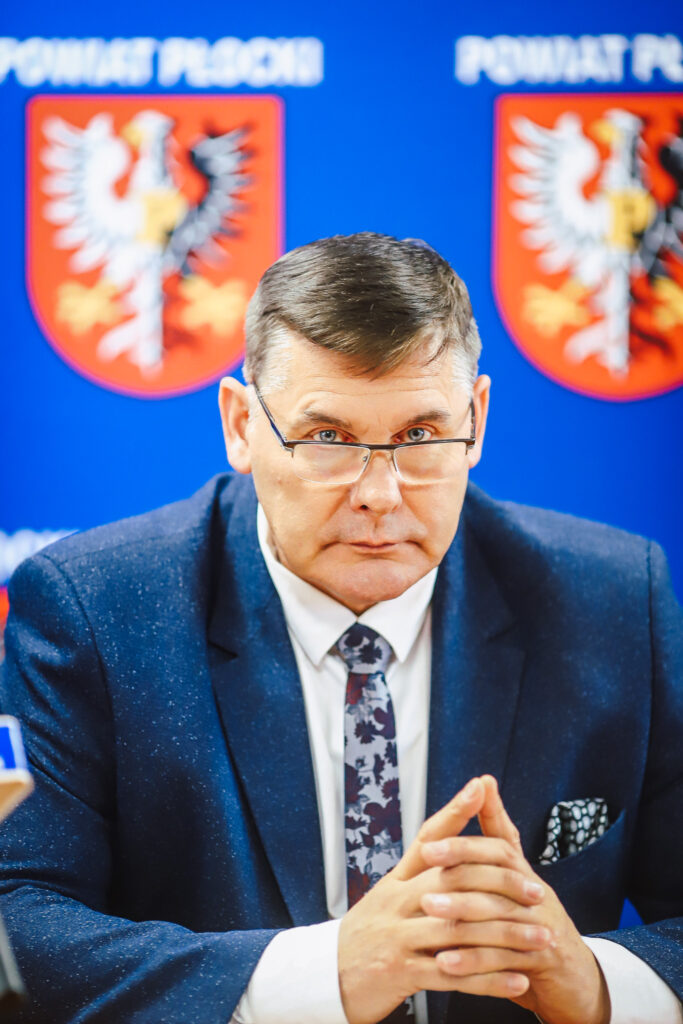 Mężczyzna w granatowym garniturze i okularach siedzi przy stole podczas noworocznego śniadania prasowego samorządowców Powiatu Płockiego, na tle niebieskiego banneru z herbem powiatu.