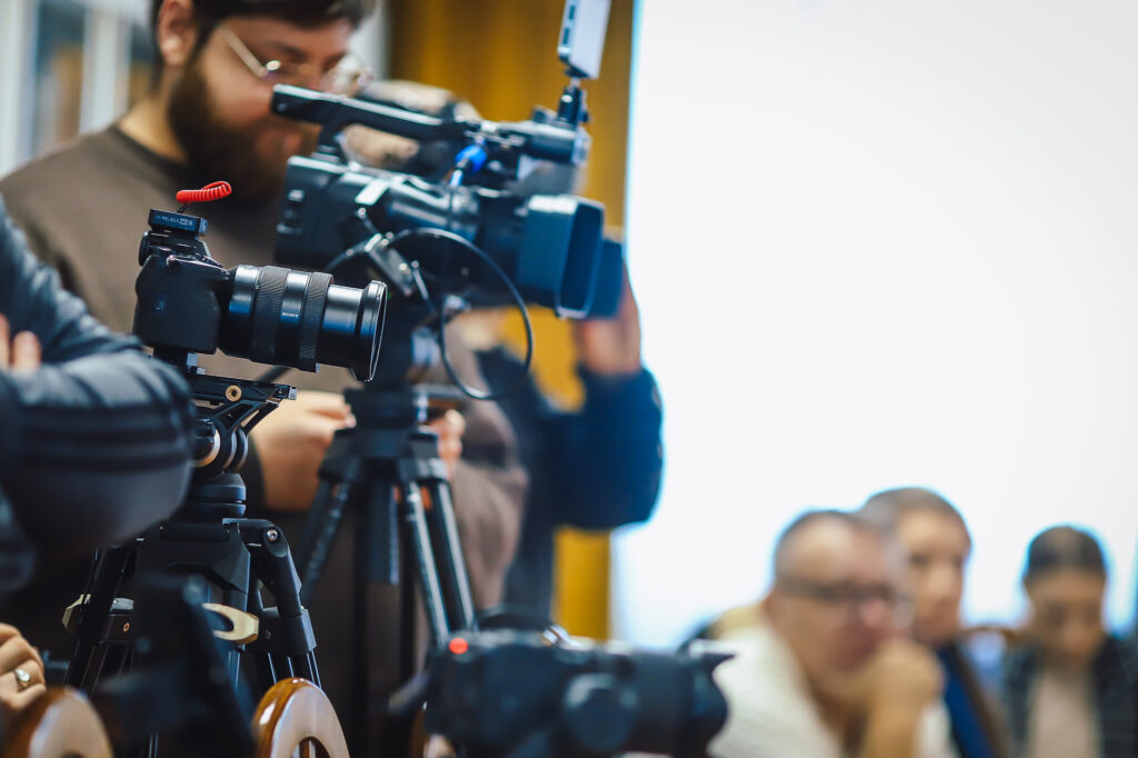 Kamera filmowa na statywie w pierwszym planie podczas noworocznego śniadania prasowego samorządowców Powiatu Płockiego z lokalnymi mediami.