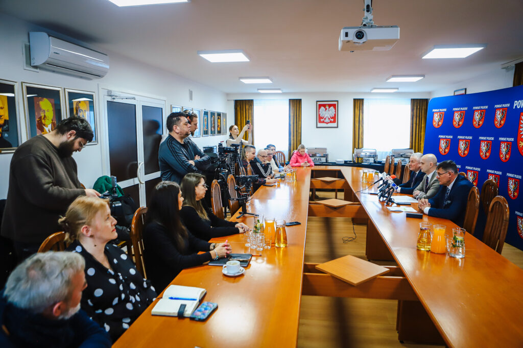 Noworoczne śniadanie prasowe samorządowców Powiatu Płockiego z lokalnymi mediami — grupa osób siedzi przy długim stole w sali konferencyjnej, z mikrofonami i kamerami, a w tle widoczny jest herb powiatu.