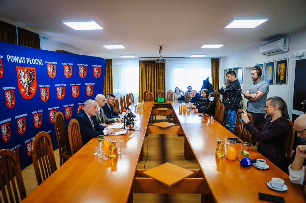 Noworoczne śniadanie prasowe samorządowców Powiatu Płockiego z lokalnymi mediami w sali konferencyjnej, przy długim stole z mikrofonami, a reporterzy i kamerzyści po prawej, na tle niebieskiej ścianki z herbem powiatu.