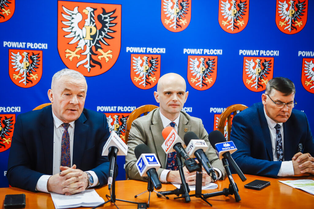 Trzech mężczyzn w garniturach siedzi przy stole z mikrofonami podczas konferencji prasowej Noworocznego śniadania samorządowców Powiatu Płockiego; w tle niebieska ścianka z herbem powiatu i napisem „Powiat Płocki”.