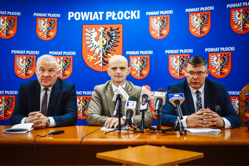 Trzech mężczyzn w garniturach siedzi przy stole podczas noworocznego śniadania prasowego samorządowców Powiatu Płockiego, za nimi niebieskie tło z herbem powiatu i napisem Powiat Płocki.