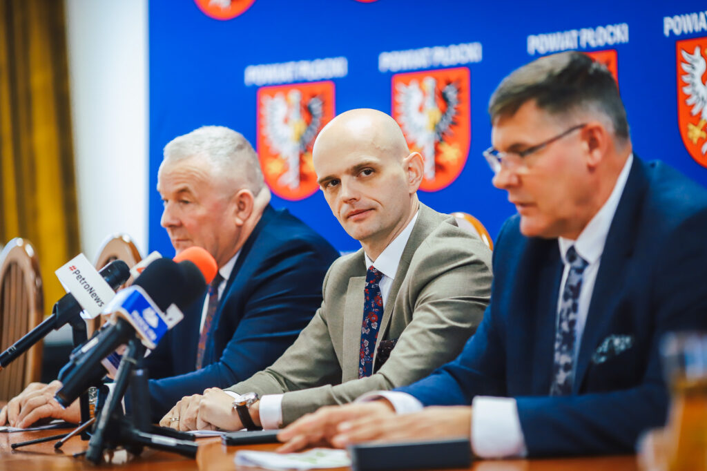 Trzech samorządowców Powiatu Płockiego podczas noworocznego śniadania prasowego z lokalnymi mediami, siedzących przy stole z mikrofonami na tle niebieskiej ściany z herbami powiatu.