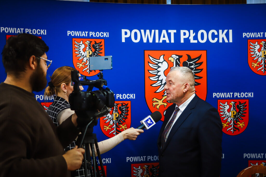 Mężczyzna w garniturze udziela wywiadu reporterce podczas noworocznego śniadania prasowego Powiatu Płockiego, na tle niebieskiej ściany z herbem i napisem Powiat Płocki.