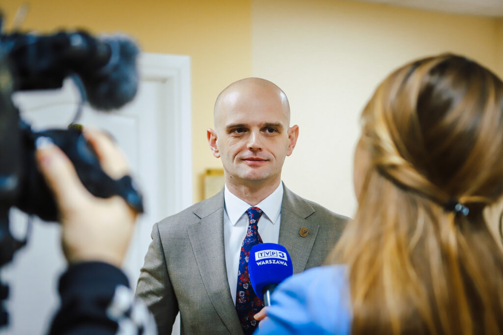 Mężczyzna w szarym garniturze udziela wywiadu reporterce z mikrofonem TVP Warszawa podczas noworocznego śniadania prasowego samorządowców Powiatu Płockiego.
